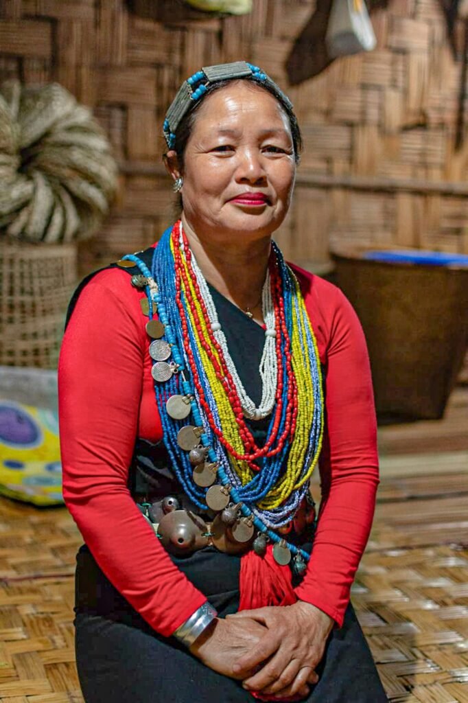 Close-up portrait of an older Naga woman from Northeast India or Myanmar wearing elaborate, stacked necklaces of blue, yellow, and white beads, large coin-like medallions, and a silver headband, set against a woven bamboo background.